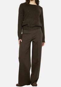 Femme portant un pull tacheté marron foncé, un pantalon large marron et des mocassins cloutés marron, sur fond blanc.