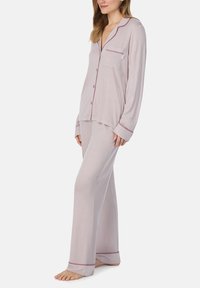 Pyjama set in licht lila stof. De top heeft een zak en contrasterende bordeauxrode piping; de broek heeft een rechte snit met gepipedde zoom.