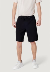 Black cotton shorts with an elastic waistband and drawstring, featuring two side pockets and a knee-length cut, worn with white sneakers.