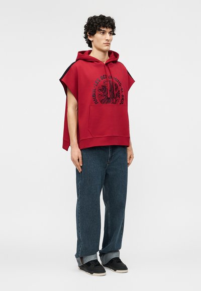 Jonge man staand, draagt een mouwloze rode hoodie met grafische tekst, losse blauwe jeans opgerold bij de pijpen, en zwarte sneakers.