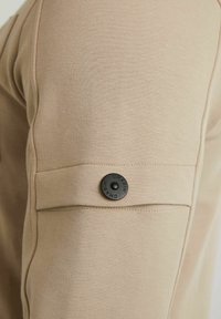Top beige a maniche lunghe realizzato in un tessuto testurizzato; presenta una chiusura a pulsante a scatto nera su una tasca laterale e leggere cuciture sui maniche.