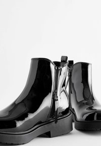 Bottines en cuir verni noir avec une finition lisse et brillante. Présentant des fermetures éclair sur les côtés, des accents texturés minimalistes, et une semelle en caoutchouc plate.