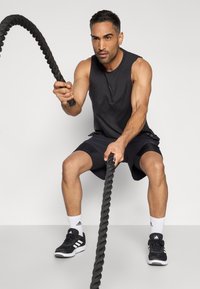 Μαύρο αθλητικό πουκάμισο χωρίς μανίκια, μαύρο σορτς, κρατώντας ένα χοντρό μαύρο σκοινί. Φορεμένο με μαύρα αθλητικά παπούτσια και λευκές κάλτσες με μαύρες λεπτομέρειες.