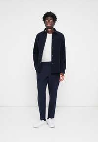 Chaqueta de pana azul marino con cierre de botón, camiseta blanca de cuello redondo, pantalones azul marino y zapatillas blancas. Presenta un diseño minimalista y un corte ajustado.