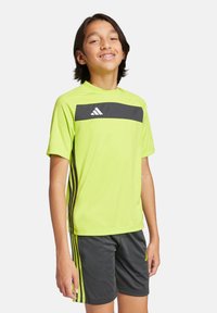 Camisa atlética amarilla brillante con una franja horizontal negra que presenta el logo de adidas; combinada con pantalones cortos gris oscuro y franjas laterales a juego.