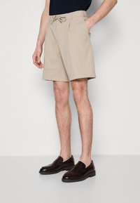 Beige shorts valmistettu kevyestä kankaasta, jossa on kiristysnauha vyötäröllä, sivutaskuja ja rento leikkaus. Yhdistetty tummiin loafereihin.
