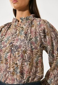 Blusa floreale con scollo arricciato, caratterizzata da un motivo a foglie multicolori in rosa, verde e oro su uno sfondo chiaro. Dettagli con bottoni visibili.