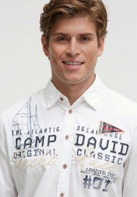 Weißes Hemd mit Knopfleiste, verziert mit nautischen Grafiken, gesticktem Text und einem kontrastierenden Aufnäher. Rote Knöpfe setzen Akzente im Design.