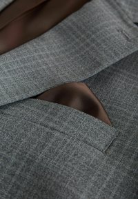 Grauer Blazerstoff mit dezentem Muster, versehen mit einem braunen Seideneinstecktuch, das aus der Brusttasche hervorlugt. Glatte Textur und maßgeschneiderte Form.