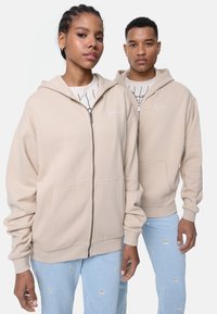 Béžová mikina na zips z jemného materiálu s prednými vreckami, rebrovanými manžetami a vyšitým logom, kombinovaná so svetlomodrými džínami s kvetinovými vzormi.