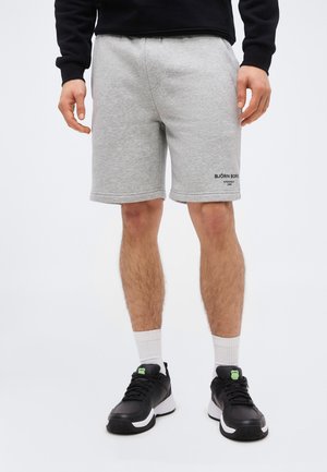 Homme portant un short Björn Borg gris clair, des baskets noires avec semelles blanches, des chaussettes blanches et un haut noir à manches longues, debout sur fond blanc.