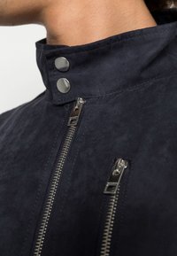 Zbliżenie osoby noszącej ciemnogranatową kurtkę z dwoma srebrnymi zamkami błyskawicznymi i stójką zapinaną na dwa metalowe zatrzaski.