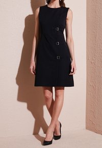 Femme portant une robe noire sans manches avec trois boucles latérales et des talons noirs, debout contre un mur beige texturé.