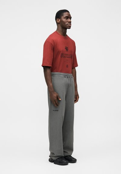 T-shirt rouge à manches courtes avec le texte "ULTRAS" et un graphisme de lion. Pantalon de survêtement gris avec cordon de serrage et logo. Baskets noires.