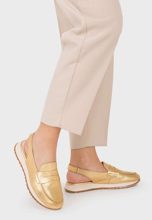 Pieds portant des mocassins slingback dorés avec un pantalon beige raccourci sur un fond clair uni.