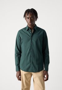 Camisa de manga larga de color verde con cuello y cierre de botones, tela texturizada y sutil detalle de logo en el pecho, combinada con pantalones beige.