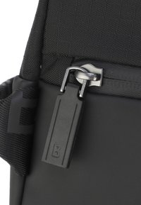 Detalle de mochila de tela negra con un tirador de cremallera plateado y una etiqueta rectangular de plástico con la letra 'B' en relieve. Se pueden ver las correas texturizadas.