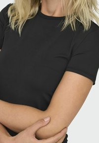 Svart kortärmad t-shirt med rund halsringning, tillverkad av ett mjukt tyg. Modellen har armarna i kors, vilket visar tröjans passform och struktur.