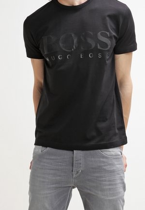 Homme portant un t-shirt noir Hugo Boss avec un grand logo en relief "BOSS" et un jean gris clair, debout devant un fond blanc.