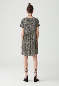 Schwarzes Blumenkleid mit kurzen Ärmeln, geraffter Taille und schwingendem Rock. Begleitet von schwarzen Knöchelsandalen mit Blockabsätzen.