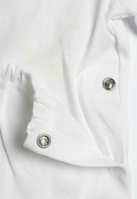 Tessuto di cotone bianco con una texture a coste. Presenta bottoni a pressione in metallo argentato all'orlo e alla manica, che consentono una facile chiusura.
