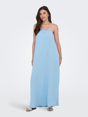 Donna che indossa un vestito lungo azzurro chiaro senza maniche con sottili spalline e sandali bianchi, in piedi su uno sfondo semplice.