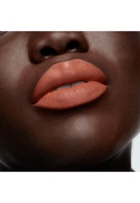 Un rouge à lèvres en terracotta mat appliqué sur des lèvres pulpeuses et lisses, sur une peau brune profonde, avec un éclat naturel et une texture des lèvres visible.