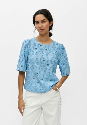 Femme portant un chemisier bleu clair texturé avec des découpes florales et un pantalon blanc, debout avec une main dans la poche sur fond blanc.