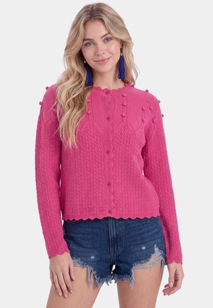 Giovane donna con lunghi capelli biondi indossa un maglione rosa lavorato a maglia con bottoni e shorts di jeans scuri consumati con orecchini blu con nappine.