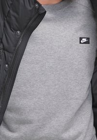 Grauer Sweatshirt mit Rundhalsausschnitt, mit einem kleinen schwarzen Nike-Logo auf der linken Brust. Teilweise von einer schwarzen Steppjacke verdeckt.