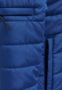 Doudoune bleue avec un design matelassé, tissu texturé, poche zippée et surpiqûres d'accentuation. Présente une surface lisse et une isolation légère.