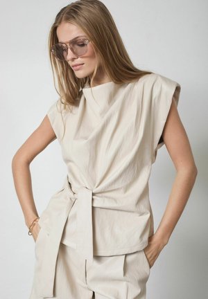 Frau mit langen Haaren, die übergroße Sonnenbrillen und ein ärmelloses beiges Oberteil mit gebundenem Gürtel sowie passende Hosen trägt, posiert mit den Händen in den Taschen.