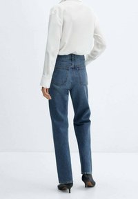 Jeans in denim blu a vita alta e gamba dritta con tasche posteriori, abbinati a una camicia bianca a maniche lunghe e scarpe nere con tacco.