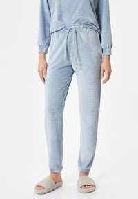 Pantalones de pijama de terciopelo suave en azul claro, con cintura ajustable y piernas ajustadas, combinados con zapatillas beige de felpa. Sin patrones visibles.