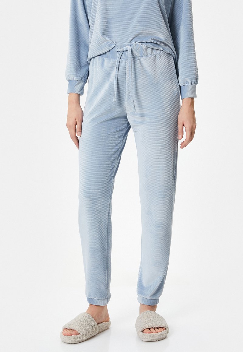 Pantalones de pijama de terciopelo suave en azul claro, con cintura ajustable y piernas ajustadas, combinados con zapatillas beige de felpa. Sin patrones visibles.