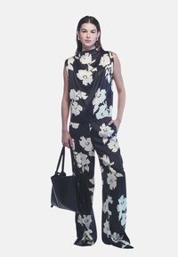 Donna che indossa un top senza maniche a fiori in bianco e nero e pantaloni a gamba larga, tiene una borsa nera con una mano mentre l'altra è in tasca.