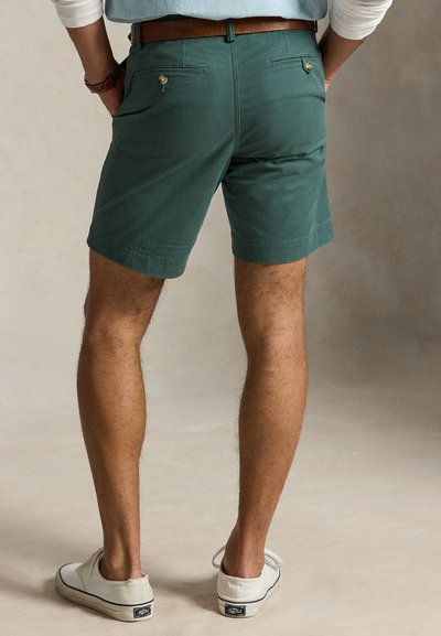 Bărbat purtând pantaloni scurți verde teal croiți pe mână, cu buzunare în spate cu nasturi, curea maro, pantofi sport albi și un top albastru deschis cu mâneci suflecate.