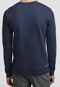 Man som bär en enkel marinblå långärmad skjorta och grå byxor, sedd bakifrån, med liten ljusblå text nära fållen.