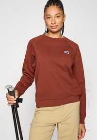 Ruda sportinė palaidinė su apvalia iškirpte ir raglano tipo rankovėmis. Ant krūtinės mažas daugiakryptis logotipas. Modelis laiko žygių lazdas.