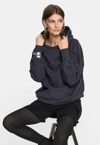 Kobieta siedząca na stołku, ubrana w luźną ciemną bluzę z kapturem, czarne szorty, czarne rajstopy, duże kółkowe kolczyki i masywną srebrną bransoletkę.