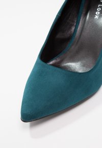Escarpin en daim bleu sarcelle avec un bout pointu et une doublure intérieure lisse. Semelle noire avec des accents minimalistes, mettant l'accent sur un design épuré.
