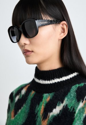 Sorte oversized solbriller med mørke glass og en teksturert svart arm, med en merket logo. Modellen har på seg en fargerik mønstrete genser.