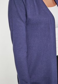 Cárdigan de punto morado con frente abierto y mangas largas, con una textura suave y un mínimo de detalles, superpuesto a una blusa blanca.