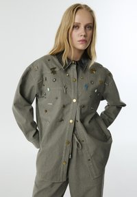 Grijze oversized jas met een zwarte kraag, versierd met verschillende metalen en emaille elementen, met een knoopsluiting en zijzakken.