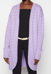 Hellblau gestrickter Cardigan mit offenem Vorderteil, strukturierten Streifen und gerippten Bündchen. Getragen über schwarzem Oberteil und Hose, die einen Gürtel hat.
