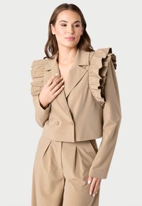 Bežinė kirpto modelio švarkas su raukšlėtomis petnešomis, dvigubomis sagomis ir lygiu paviršiumi. Derinama su atitinkančiomis aukštu liemeniu kelnėmis.