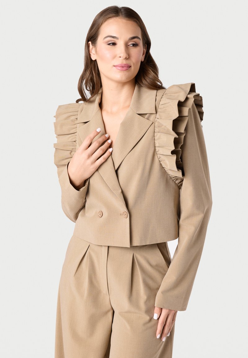 Bežinė kirpto modelio švarkas su raukšlėtomis petnešomis, dvigubomis sagomis ir lygiu paviršiumi. Derinama su atitinkančiomis aukštu liemeniu kelnėmis.