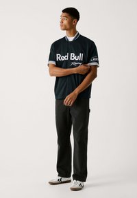 Dunkelblaues Kurzarm-Shirt mit "Red Bull Racing"-Logo, weißen Streifenakzenten am Kragen und an den Ärmeln, kombiniert mit schwarzen Hosen und weißen Schuhen.