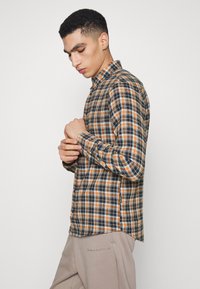 Chemise à carreaux en bleu marine, orange et blanc. Elle présente des manches longues, un col et une fente sur le côté, fabriquée en tissu léger.