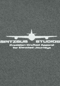 Grijze stof met witte bedrukt tekst en de silhouet van een vliegtuig. De tekst luidt "SPITZBUB STUDIOS" en "Nauwkeurig vervaardigde kleding voor verhoogde reizen."